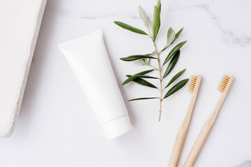 Top view of white plastic tube without label, bamboo toothbrushes and olive branch on white...