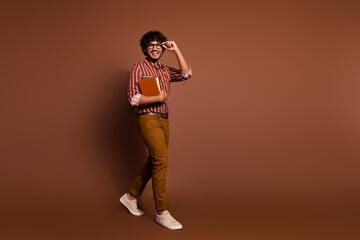 Full body portrait of nice young man hold book walk empty space wear striped shirt isolated on brown color background
