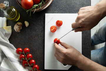 Male hands cutting cherry tomatoes at home. Fresh tomatoes in bowl, olive oil, rosemary and marble...
