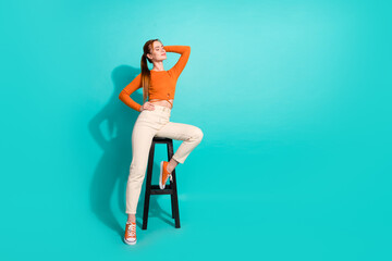 Full length photo of lovely woman wear crop top white pants sit on chair hold arm on head posing isolated on turquoise color background