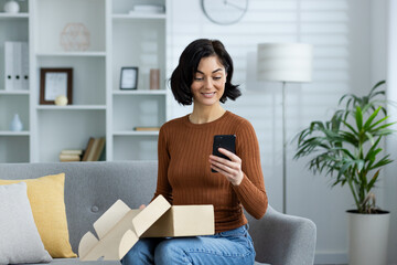 Happy woman unboxing package and using smartphone while sitting on sofa at home