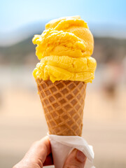Refreshing orange sorbet in an ice cream cone held by a hand with defocused beach background,...