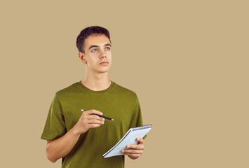 Portrait of thinking brunette boy holding pen and notepad in hands isolated on beige studio...
