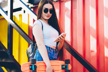 Stylish hipster girl in sunglasses and skateboard in hands standing on stairs and making payment...