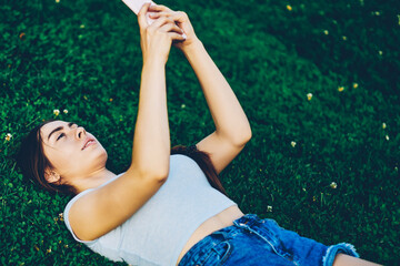 Attractive young woman with braid lying on green lawn and chatting online with friends on smartphone device connected to 4G internet.Hipster girl enjoying summer time on grass and making selfie photo
