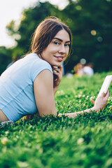 Portrait of attractive smiling young woman lying on green lawn in park and looking at camera while sharing media in social networks on smartphone.Cheerful blogger publishing post on cellular