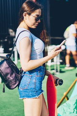 Side view of attractive female lover of skateboarding reading incoming notification on smartphone.Charming hipster girl in sunglasses and stylish backpack updating media on telephone standing outdoors