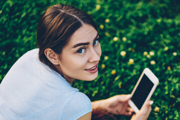 Portrait of beautiful brunette hipster girl smiling at camera while installing mobile application for editing photos on smartphone lying on green lawn in park.Charming cute blogger publishing post