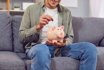 Man sits on a couch at home, putting a coin into a piggy bank. He is happy and focused, planning...