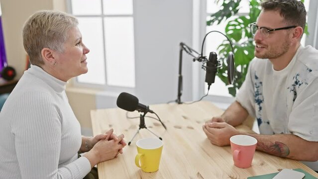 A woman podcaster interviews a man in a bright indoor studio setup with microphones and coffee mugs on the table.