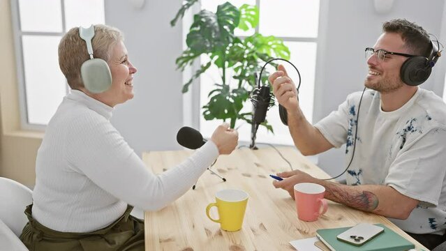 A mature woman and a young man fist bumping in a bright radio studio during an engaging podcast interview.