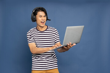 Happy young Asian pro gamer with headphones holding computer laptop and win in online video game isolated over blue background, Cyber sportsman playing tournaments concept