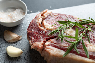 Close-up of fresh raw beef steak garnished with rosemary on cutting board. Salt and garlic cloves on kitchen counter