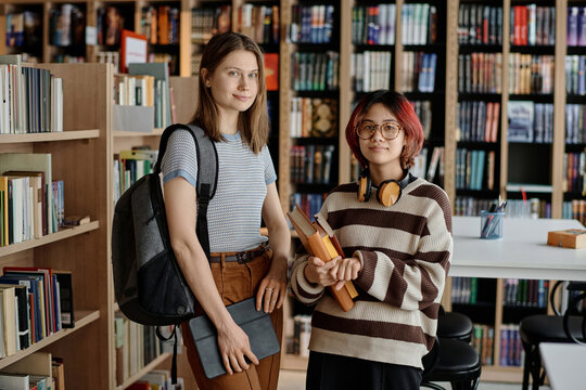 Medium long shot of two Asian and Caucasian female students wearing casual clothes standing in library looking at camera