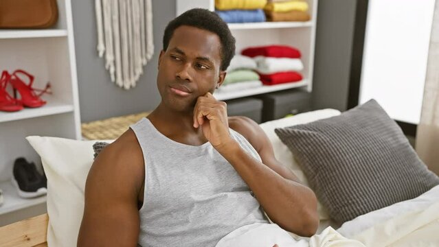 Thoughtful young african american man unsure in his pyjamas, hand on chin, expressing doubt in bedroom - question mark hovering above his pensive face
