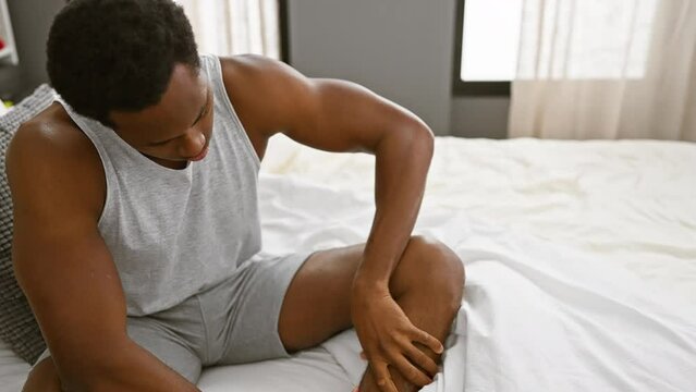 Adult black man experiencing leg pain while sitting on a bedroom bed indoors