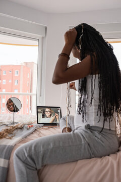 Happy woman making braids using laptop on bed at home