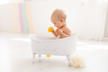 a little baby girl of six months with blue eyes bathes in a bubble bath and plays with rubber toys...