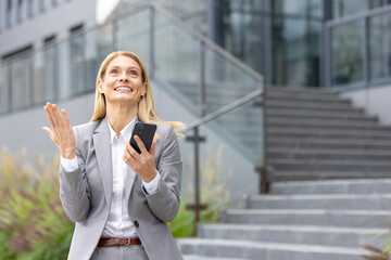 An accomplished businesswoman in a gray suit radiates happiness and selfassurance while celebrating outside a sleek office building, holding a mobile phone, standing on the steps