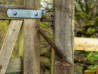 Close-up of an old wooden gate with rusty spring and hinge mechanism, surrounded by lush greenery in a rural outdoor setting.