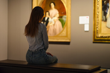 Thoughtful young Caucasian woman wearing glasses and looking at exhibition. Concept of Museum Day.