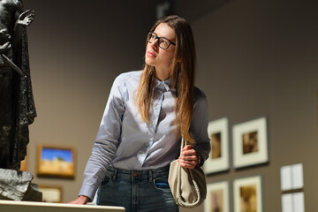 Thoughtful young Caucasian woman wearing glasses and looking at exhibition. Concept of Museum Day.