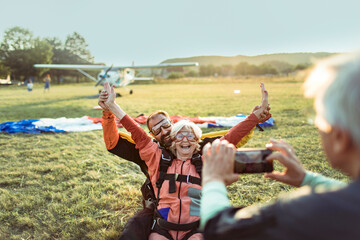 Senior woman celebrating after a successful skydive land with instructor with husband capturing the moment