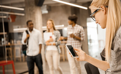 Businesswoman using smartphone in modern office with colleagues in background
