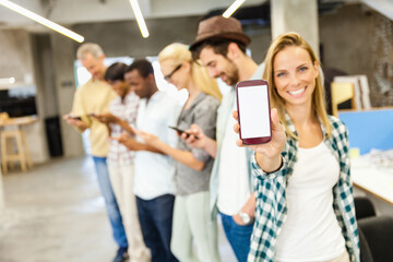 Smiling young woman showing smartphone with blank screen with diverse colleagues in background at office