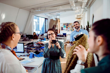 Smiling female fashion designer holding a camera in a lively design studio with colleagues...
