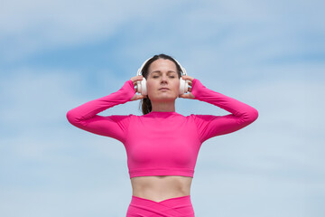 Sporty woman listening to music with headphones, wearing pink sports clothing against a blue sky
