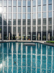 modern building with a facade of large glass windows reflecting the surrounding architecture. In front of the building is a pool with clear blue water, creating reflections of the windows