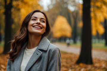 Perfect smiling lady in grey coat posing in autumn park, looking up away. Portrait of happy young woman enjoying on fresh air on September fall day. Leisure activity concept. Copy ad text space