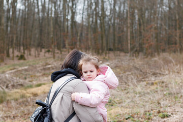 A little cute girl in the arms of her mother walking with a backpack through a snowless winter forest