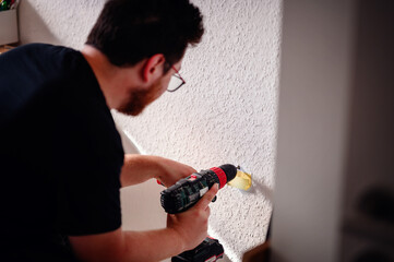 A man with glasses and a beard uses a power drill to drill into a wall, focused on the task as part...