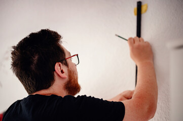 A man with glasses and a beard marks the wall with a pencil for shelf installation, focusing on...