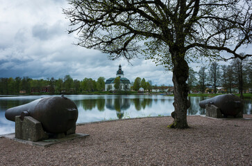 Filipstad's Church and cannons on Kanonudden in Filipstad, White church building in the distance is reflecting in the water, Two large cannons, A dark tree with small leaves, Cloudy weather