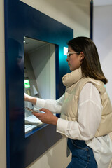 A woman wearing glasses and a light-colored vest uses an ATM machine at a bank, holding her smartphone in one hand in an indoor setting.