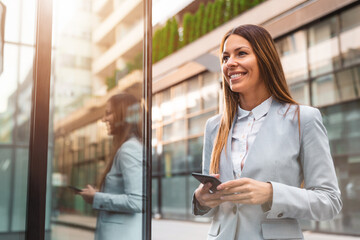 Smiling businesswoman using mobile phone while walking by the office building.