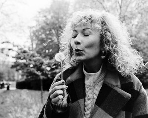 Monochromatic image of curly woman blowing a dandelion in a park.