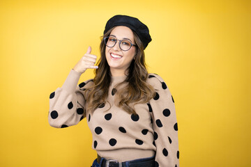 Young beautiful brunette woman wearing french beret and glasses over yellow background smiling doing phone gesture with hand and fingers like talking on the telephone