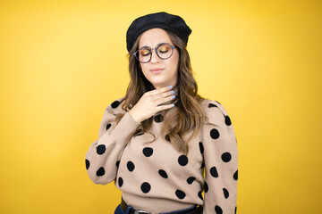 Young beautiful brunette woman wearing french beret and glasses over yellow background touching...