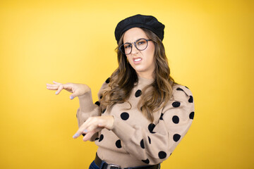 Young beautiful brunette woman wearing french beret and glasses over yellow background disgusted...