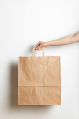 Woman’s hand holding craft paper bag against a white wall. Paper bag  mock up for your design;  ...