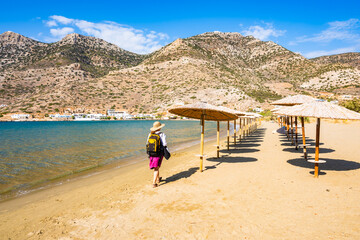Unidentified woman tourist wearing sun hat and backpack walking on sandy beach with crystal clear...