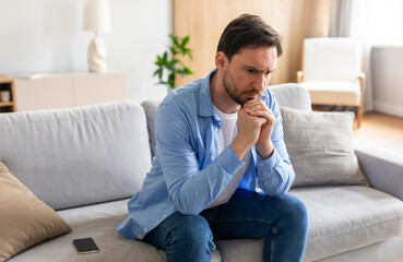 A man is seated on a couch, deep in thought, with his hand resting on his chin. He appears contemplative and engaged in introspection.