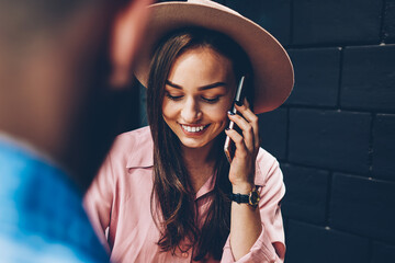 Cheerful trendy dressed woman talking on cellular during meeting with boyfriend in cafe, hipster girl having telephone conversation about booking satisfied with communication spending time with friend