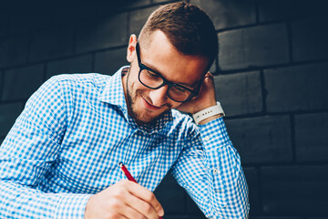 Positive hipster guy in spectacles concentrated on writing ideas for coursework project in college campus,skilled male architect drawing graphic in sketchbook satisfied with creative working process
