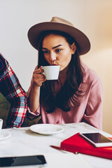 Young male and female friends resting in cafe interior drinking coffee during work break, trendy dressed woman in hat spending time with her boyfriend in eyewear thinking about plans for leisure