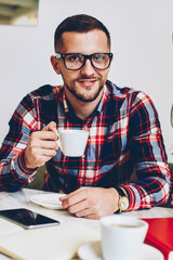 Young woman in trendy hat talking on phone while drinking coffee with her boyfriend in cafe, handsome man in eyewear looking at camera having meeting with female colleague making telephone call